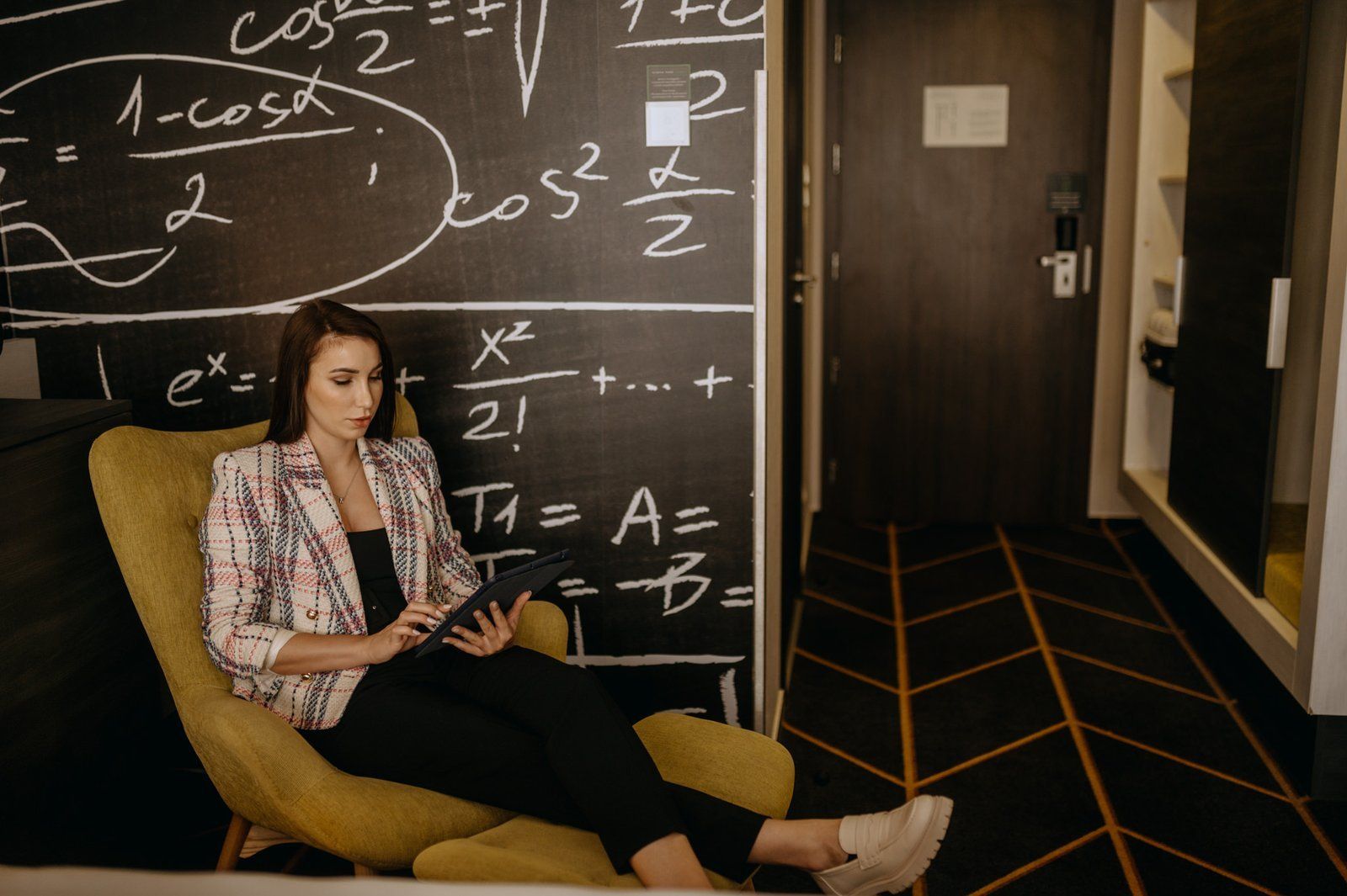 Un oaspete citește într-un fotoliu în cameră; pe perete, o tablă de cretă plină de formule matematice
