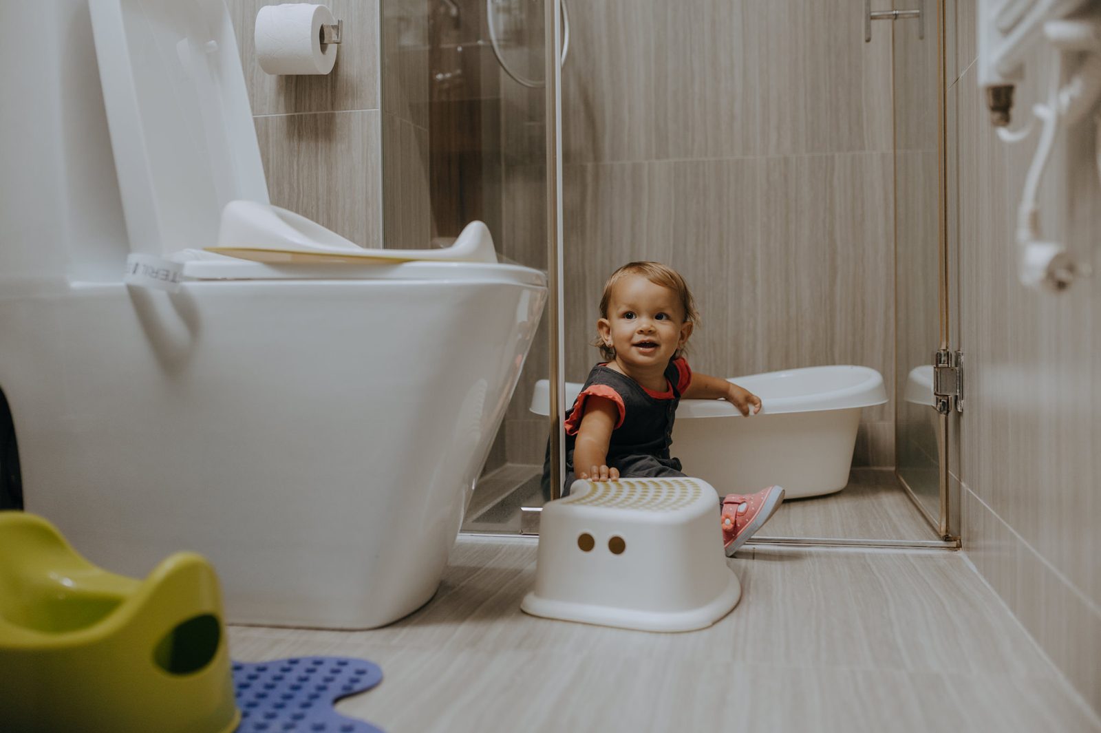 Copil mic în baie, lângă o treaptă, o oliță și un reductor de WC